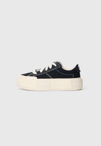 Canvas sneaker with a black upper, white stitching, and a thick cream rubber sole. Features flat laces and a tag on the back.