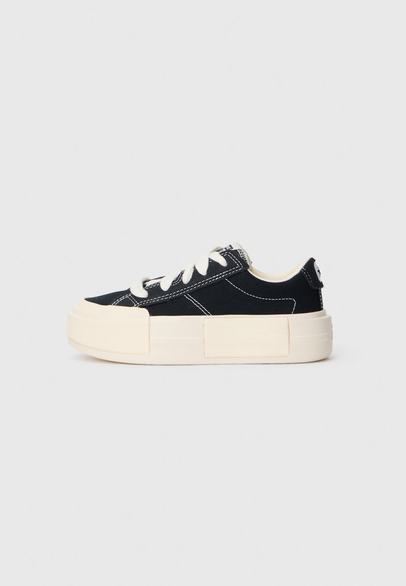 Canvas sneaker with a black upper, white stitching, and a thick cream rubber sole. Features flat laces and a tag on the back.