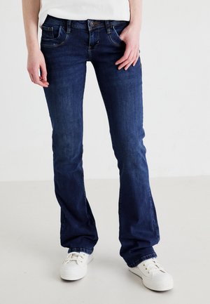 Osoba ubrana w ciemnoniebieskie jeansy typu bootcut oraz białe trampki, stojąca z rękami swobodnie opuszczonymi wzdłuż ciała, na jednolitym jasnym tle.