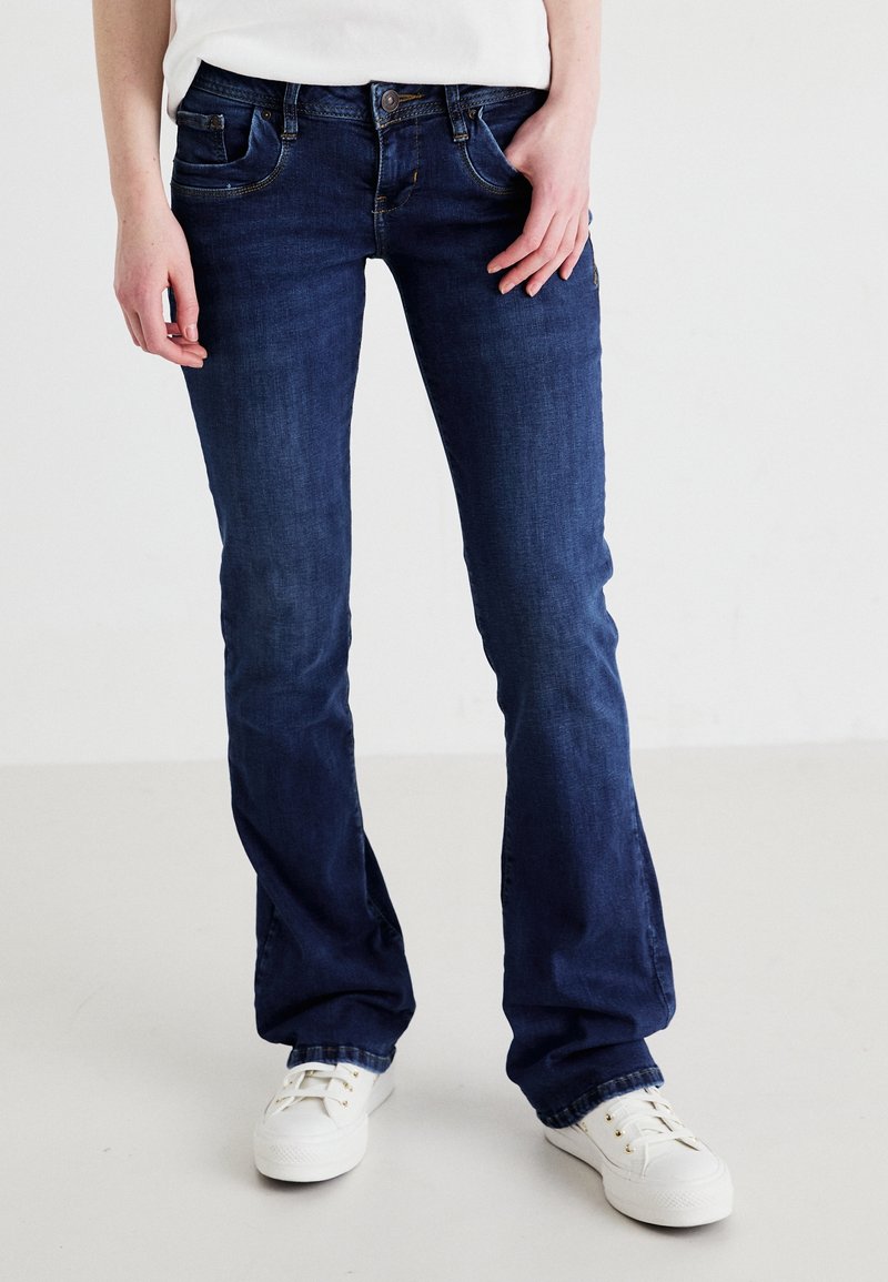 Personne portant un jean bootcut bleu foncé et des baskets blanches, debout, les mains détendues le long du corps sur un fond clair uni.