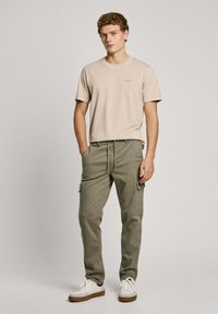 Camiseta de algodón beige de manga corta, combinada con pantalones cargo verdes oliva con bolsillos laterales. Zapatillas blancas con suelas marrones.