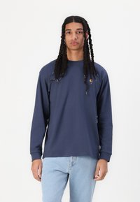 Long-sleeve navy shirt in soft fabric with a crew neckline and side logo. Paired with light blue jeans for a casual look.