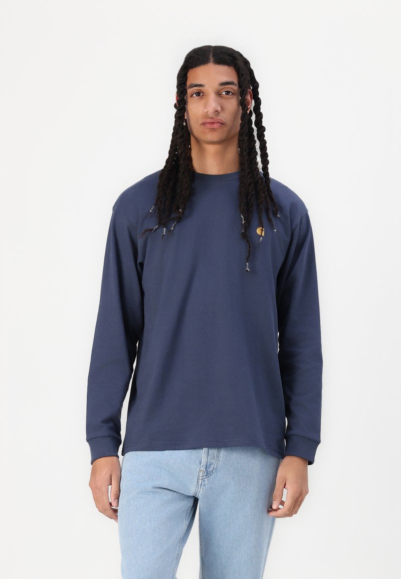 Long-sleeve navy shirt in soft fabric with a crew neckline and side logo. Paired with light blue jeans for a casual look.