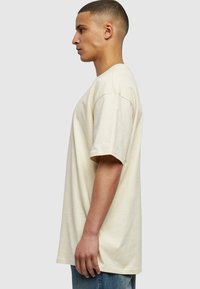 Lys beige oversized t-skjorte, kort erm, laget av myk bomull, med rund hals og avslappet passform. Brukt sammen med blå denimjeans.
