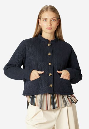 Marineblauwe gewatteerde jas met hoge kraag, voorzien van gouden knopen en voorzakken, gedragen over een meerkleurige gestreepte blouse.