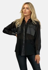 Czarna przezroczysta koszula zapinana na guziki z kontrastującymi, gładkimi kieszeniami, ozdobionymi ćwiekami na rękawach oraz gładką fakturą. Połączona z czarnymi legginsami.