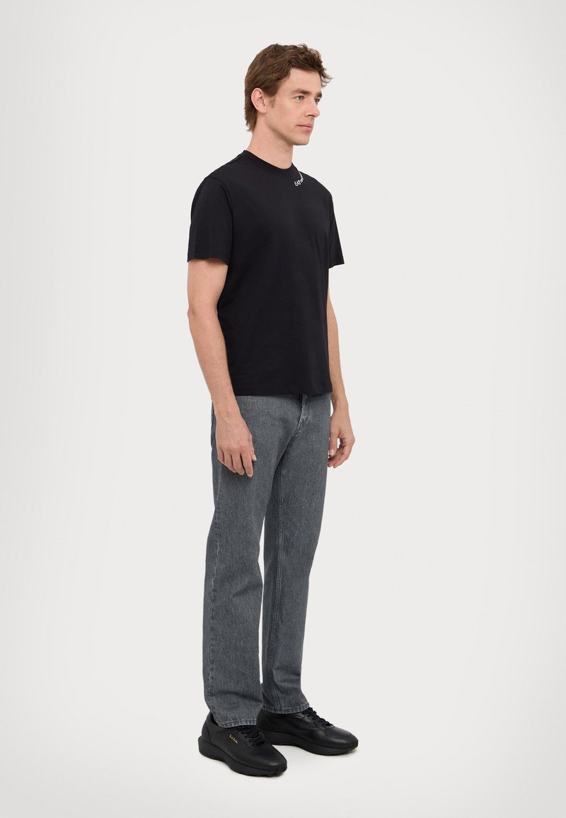 T-shirt in cotone nero, collo rotondo, design semplice; abbinato a jeans grigi e sneakers nere con dettagli dorati. Outfit semplice e casual.