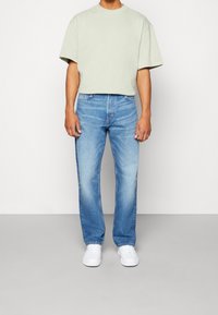 T-shirt corta verde chiaro con spalle cadute, abbinata a jeans dritti blu classici e sneakers bianche. Texture in cotone morbido.