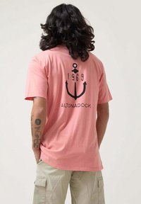 Camiseta de color rosa claro con un gráfico de ancla negra, "1969" y "ALTONADOCK" impresos en la parte de atrás; ajuste casual y material suave.