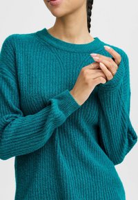 Pull en maille côtelée teal avec un col rond et des manches longues, fabriqué à partir d'un tissu en tricot texturé. Les accents comprennent des poignets et des motifs subtils.