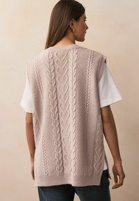 Vesta tricotată roz deschis, cu un model de tricotaj în formă de cablu, având un finisaj texturat, decolteu rotund și fante laterale, purtată peste o cămașă albă.