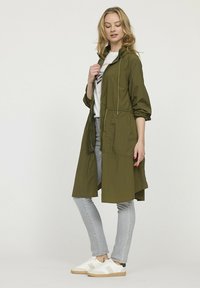 Manteau léger de couleur vert olive avec une fermeture éclair à l'avant, une taille élastique, des poches latérales et une coupe décontractée, associé à un jean clair et des chaussures blanches.