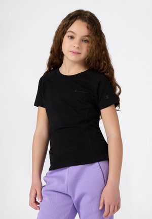 Jeune fille avec de longs cheveux bouclés portant un t-shirt Champion noir et un pantalon violet clair, se tenant devant un fond blanc uni.