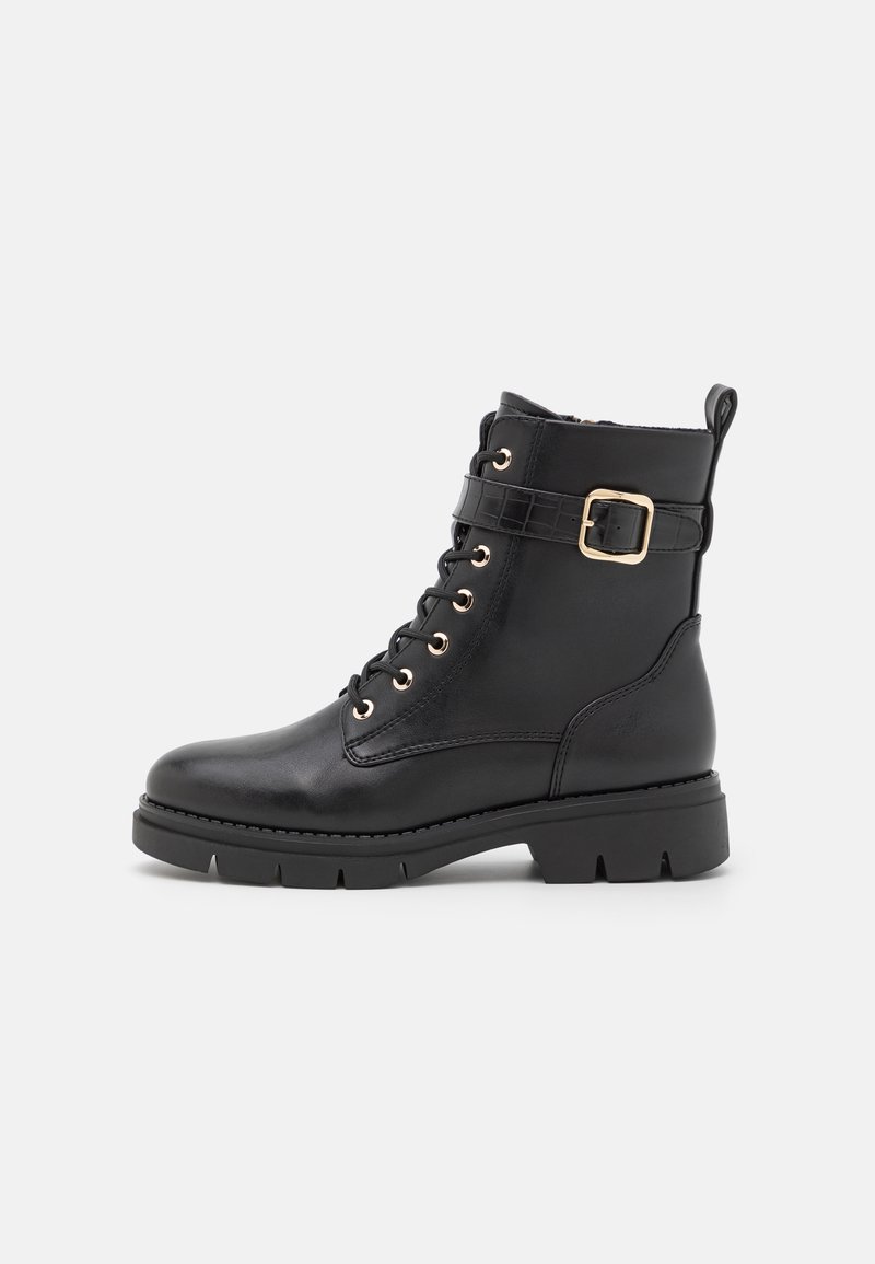 Bottine en cuir noir avec une semelle plate, œillets dorés, laçage frontal et un accent de boucle sur une sangle texturée autour de la cheville.