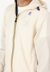 Světle krémová pláštěnka z hladké, voděodolné látky, vybavená zipem s barevnými pruhy a kapucí.