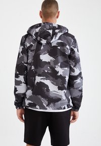 Schwarz-graue, abstrakt gemusterte Jacke mit Kapuze, aus leichtem Material; verfügt über elastische Bündchen und einen dezenten Glanz.