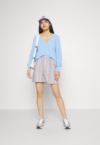 Top azul claro de mangas compridas e decote em V, calções às riscas multicoloridos, ténis cinzentos e um chapéu claro, com uma pequena mala branca. Fundo plano.