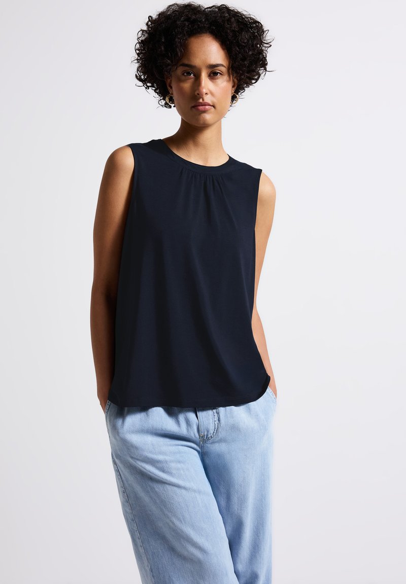 Top sans manches bleu marine avec un col rond et des détails plissés, assorti à un pantalon en denim bleu clair, ample. Texture de tissu lisse.