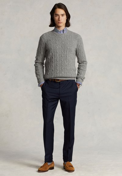 Man standing with hands in pockets wearing a gray cable knit sweater, striped shirt, navy trousers, and brown suede tassel loafers.