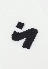 Biała skarpetka z wypukłym czarnym logo. Logo ma charakterystyczny kształt z wyraźnymi liniami i kwadratowym akcentem, teksturowanym w stosunku do tkaniny.