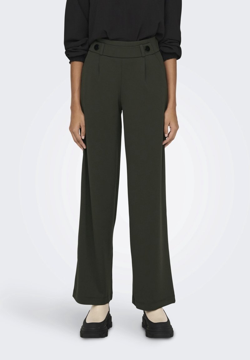 Pantalons larges en tissu vert foncé, dotés d'une taille haute avec deux grands boutons et d'une texture lisse. Portés avec des chaussures noires.