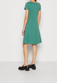 Zielona sukienka z krótkim rękawem z dopasowanym gorsetem i rozkloszowanym dołem, wykonana z miękkiego materiału; w połączeniu z czarnymi butami na platformie.
