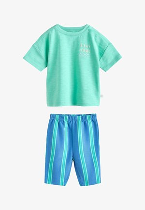 Kisgyermek rövid ujjú halványzöld ingben, amelyen a "Maradj hűvös" felirat áll, kék nadrágban, melyen függőleges türkiz csíkok láthatók.