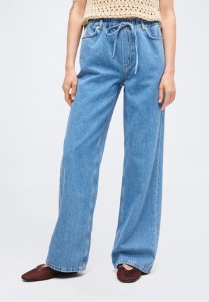 Γυναίκα που φοράει ανοιχτό μπλε τζιν φαρδιά στο κάτω μέρος με κορδόνι στη μέση και μπορντό επίπεδα παπούτσια, στέκεται μπροστά σε απλό φόντο.