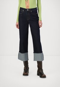 Jeans de mezclilla anchos de color azul oscuro con puños de mezclilla azul claro. Presentan bolsillos frontales y costuras en contraste. Combinados con botas marrones hasta la rodilla.