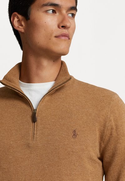 Zip-up tröja i ljusbrunt stickat tyg, med ribbat krage och en liten broderad logotyp på bröstet.