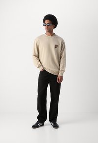 Felpa beige con scollo rotondo e logo dell'orso, abbinata a pantaloni neri e scarpe da ginnastica nere con strisce bianche. Il modello indossa occhiali da sole scuri.