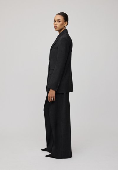 Completo nero su misura in vestibilità slim, caratterizzato da un lungo blazer e pantaloni a gamba larga. Tessuto liscio con dettagli minima e linee decise.