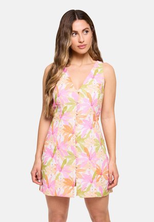 Mujer con cabello largo y ondulado que lleva un vestido sin mangas con botones delante con un patrón floral en tonos rosa, naranja y verde sobre un fondo claro.