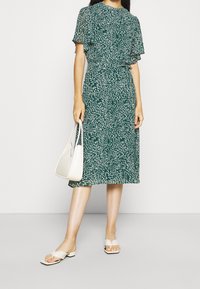 Grön blommig midiklänning med korta, svävande ärmar, åtdragen midja och flödande kjol. Kombineras med vita slidesandaler och en väska.