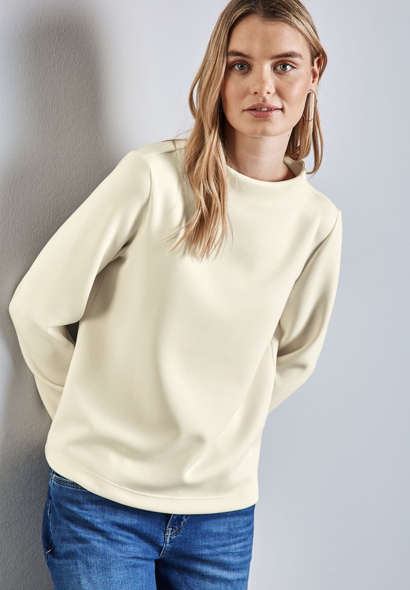 Top a maniche lunghe color crema con scollatura rotonda, texture liscia, vestibilità morbida e spalle scese, abbinato a jeans blu.