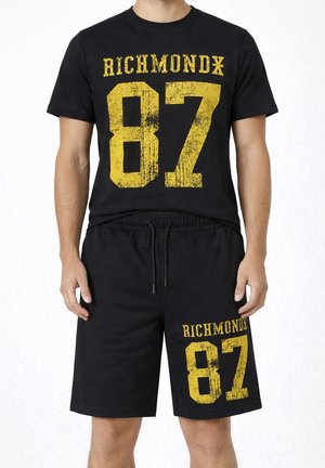 Uomo che indossa una maglietta nera a maniche corte e pantaloncini con grande stampa gialla "RICHMOND 87" sul petto e sulla gamba sinistra.
