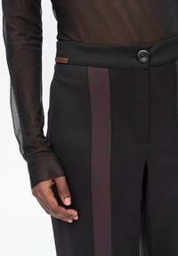 Czarne spodnie z ciemnymi pionowymi paskami po bokach noszone z przezroczystą bluzką z długim rękawem, pokazujące ciemnoskórą dłoń spoczywającą przy boku.