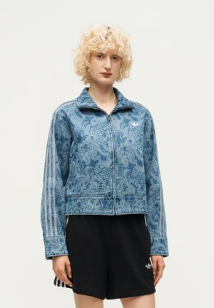 Person mit kurzen lockigen blonden Haaren, die eine blaue gemusterte Reißverschlussjacke und schwarze Shorts mit Adidas-Logo trägt, steht vor einem schlichten Hintergrund.