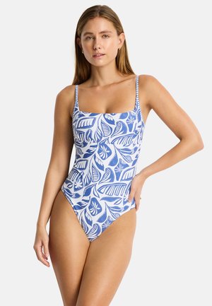 Kobieta ubrana w jednoczęściowy kostium kąpielowy z niebiesko-białym tropikalnym wzorem liści, stojąca z jedną ręką na biodrze na jednolitym tle.
