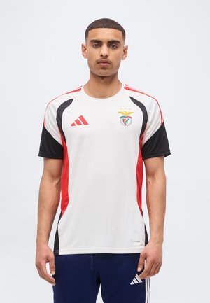 Mladý muž oblečený v bielom Adidas športovom drese s červenými a čiernymi akcentmi a logom futbalového klubu Benfica, stojaci pred jednoduchým pozadím.