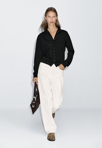 Schwarzes Hemd mit langen Ärmeln, kombiniert mit weißen weit geschnittenen Hosen und einer braunen Handtasche. Das Model trägt braune Sandalen. Einfaches Design.