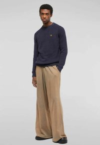 Giovane uomo che indossa un maglione blu navy e pantaloni beige larghi, in piedi con una mano in tasca su uno sfondo semplice.