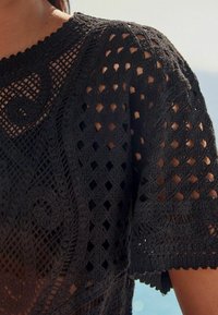 Černý háčkovaný top s otevřeným mřížkovým designem, který má diamantové tvary a vířivou dekoraci. Texturovaný povrch s vroubkovaným výstřihem a rukávy.