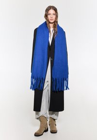 Blå frynset uldhalstørklæde hængende over en sort frakke, parret med grå bukser og brune ankelstøvler i et minimalistisk miljø.