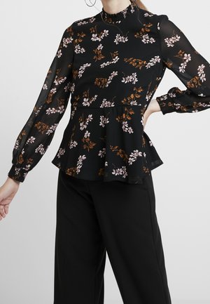 Femme portant un chemisier noir transparent à motifs floraux avec de longues manches bouffantes et un pantalon noir, posant avec une main sur la hanche et l'autre tendue.