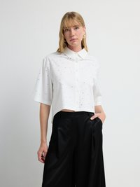 Hvid cropped skjorte med krave, prydet med spredte sølvdekorationer. Parret med sorte plisserede bukser med brede ben, glat tekstur.