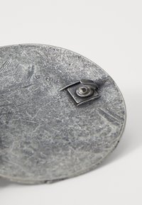 Apaļa metāla plāksne ar teksturētu virsmu, kurai ir neliels kvadrāta stiprinājums un apaļa skrūve. Metāliska pelēka krāsa un nolietots apdare.