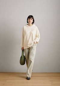 Beige φούτερ με λογότυπο στο στήθος, συνδυασμένο με χαλαρούς, ανοιχτούς πράσινους παντελόνες. Το μοντέλο κρατάει μια πράσινη τσάντα και φοράει παπούτσια με μαύρο σχέδιο.