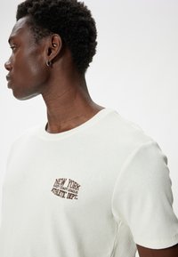 Lys beige ribbet T-shirt med et brunt broderet design "NEW YORK EAST COAST LEAGUE ATHLETIC DEPT." på venstre bryst.