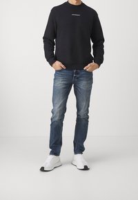 Musta pyöreäkauluksinen collegepaita, jossa valkoinen logo, yhdistetty vaaleansinisiin kuluneisiin farkkuihin ja valkoisiin tennareihin, joissa on mustia yksityiskohtia.